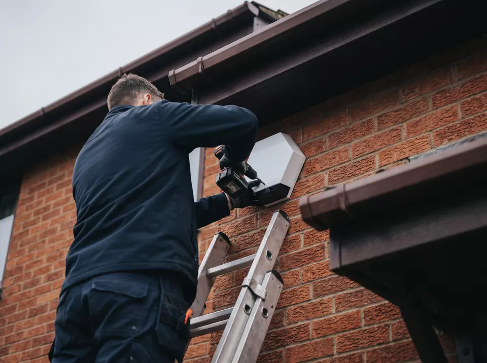 An alarm engineer on a ladder fixing a bell box to a red-brick house wall