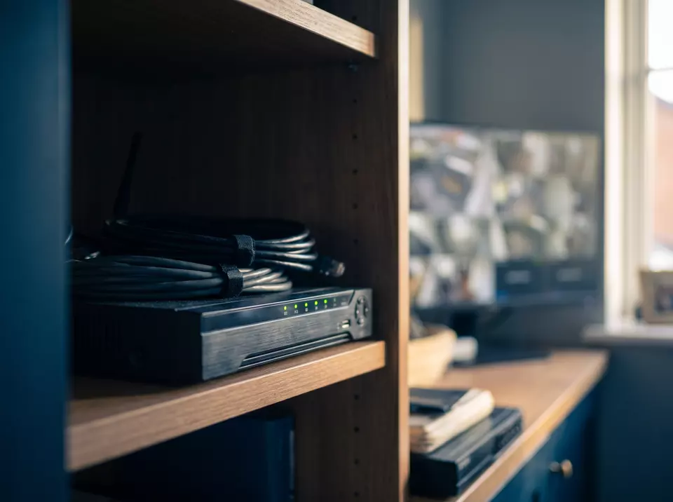 A CCTV recorder on a shelf with network cables and a monitor showing camera views behind