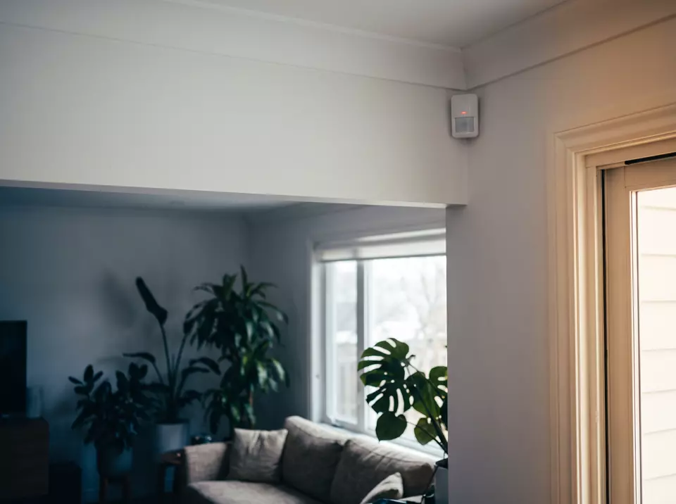 A motion sensor in the corner of a living room ceiling with its indicator light on