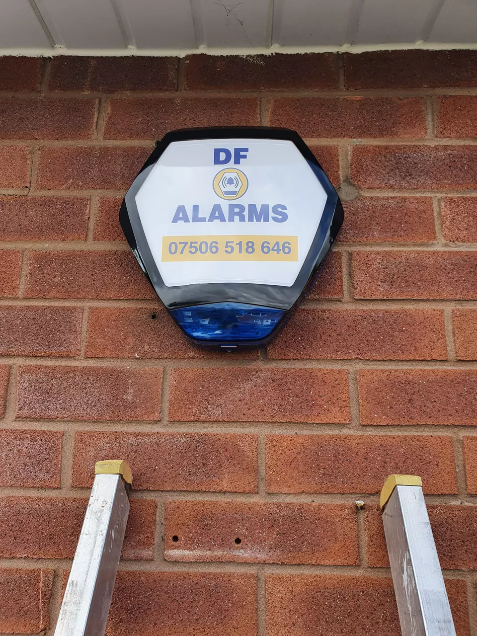 A DF Alarms bell box above a ladder on a red-brick wall